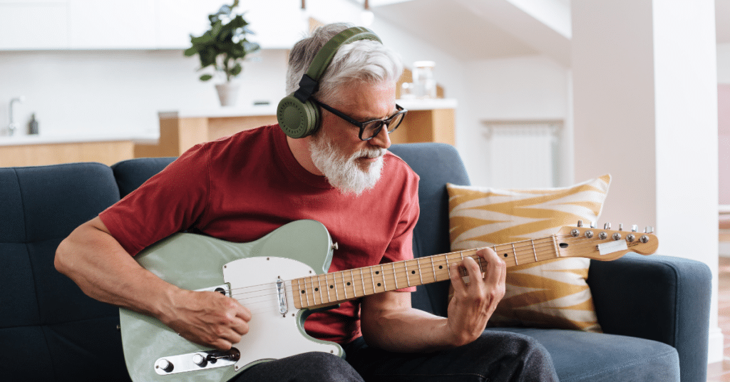 Mann mit E-Gitarre und Kopfhörern lernt online im Wohnzimmer vor dem Laptop – Rock & Pop Gitarrenkurs der New Music Academy.
